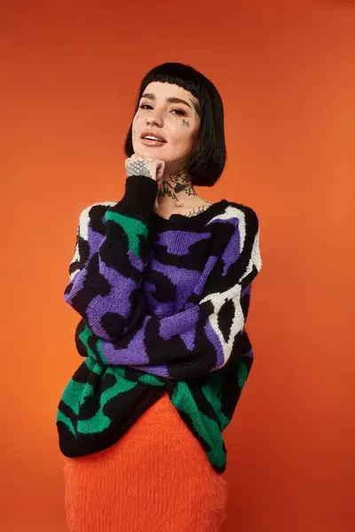 A young woman with short hair and unique tattoos showcases her style in a bright sweater against an orange background. — Stock Photo