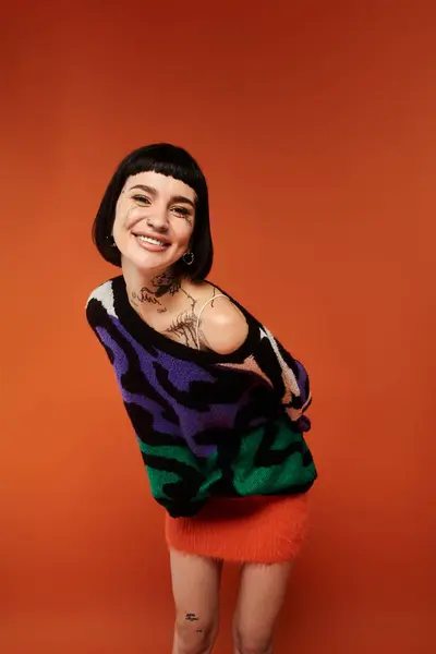A young woman with short hair and tattoos joyfully poses in a colorful sweater against a vivid orange backdrop. — Stock Photo