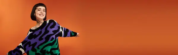 Una joven con el pelo corto y tatuajes posa alegremente en un suéter colorido sobre un fondo naranja brillante. - foto de stock