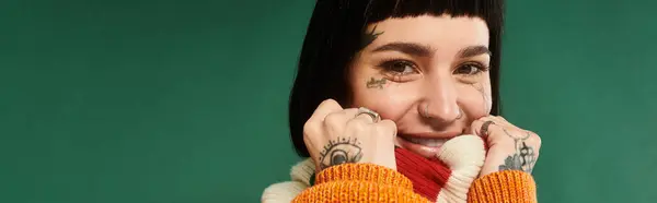 A young woman with tattoos and short hair playfully poses in a vibrant sweater against a rich green background. — Stock Photo