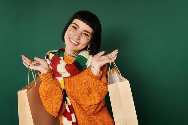 Une jeune femme joyeuse dans un pull chaud tient des sacs à provisions, souriant avec confiance dans un cadre de studio confortable. — Photo de stock