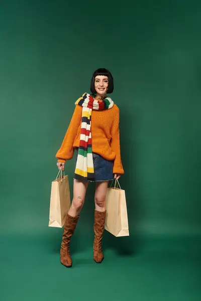 A young woman showcases her unique style wearing a warm sweater and holding shopping bags in a studio. — Stock Photo