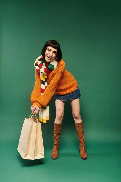 A young woman with short hair and tattoos smiles while wearing a warm sweater and holding shopping bags. — Stock Photo