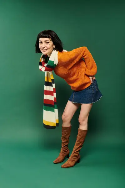 A cheerful young woman with short hair and tattoos poses playfully in a cozy sweater against a green backdrop. — Stock Photo