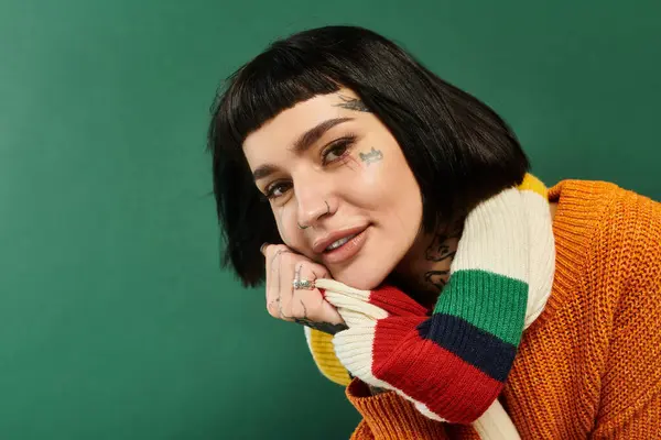 Una joven con una cálida sonrisa exhibe sus tatuajes y suéter acogedor en un ambiente de estudio con un fondo verde. - foto de stock