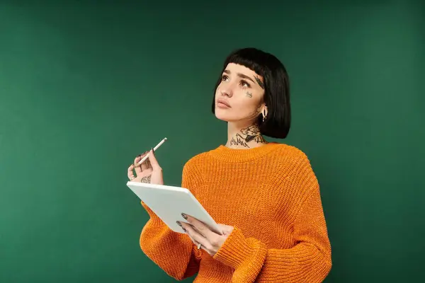 Una mujer joven con el pelo corto y tatuajes cuidadosamente sostiene una tableta mientras usa un suéter cálido y acogedor. — Stock Photo