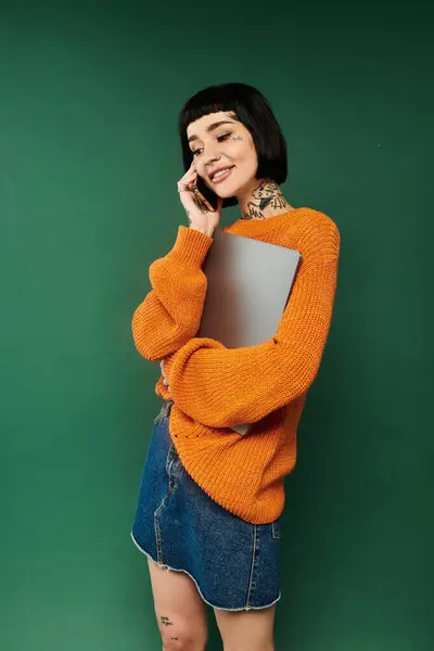 A young woman with short hair and tattoos beams as she holds a laptop in a warm, cozy sweater. — Stock Photo