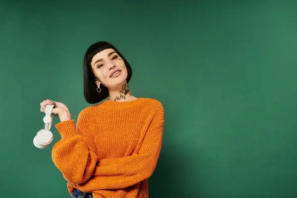 A young woman with short hair and tattoos smiles warmly while holding headphones, dressed in a comfy sweater. — Stock Photo