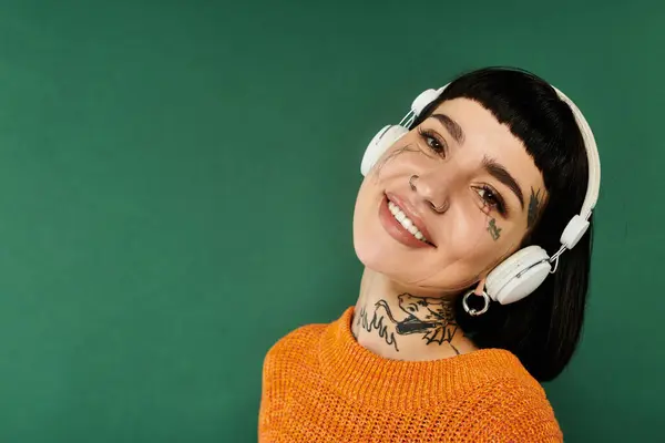 A young woman with short hair and tattoos smiles warmly while wearing a cozy sweater and headphones. — Stock Photo