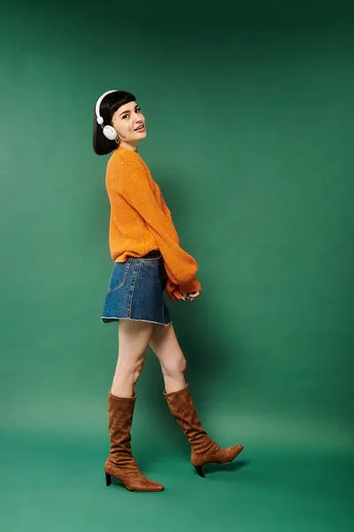 A young woman with short hair and tattoos stands confidently in a cozy sweater on a green backdrop. — Stock Photo