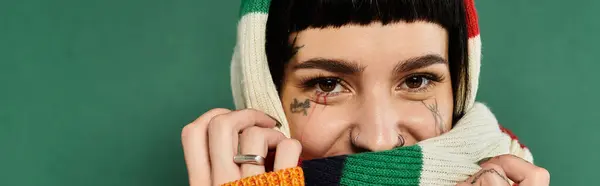 A young woman playfully poses with a bright sweater and scarf, showcasing her tattoos against a warm green backdrop. — Stock Photo