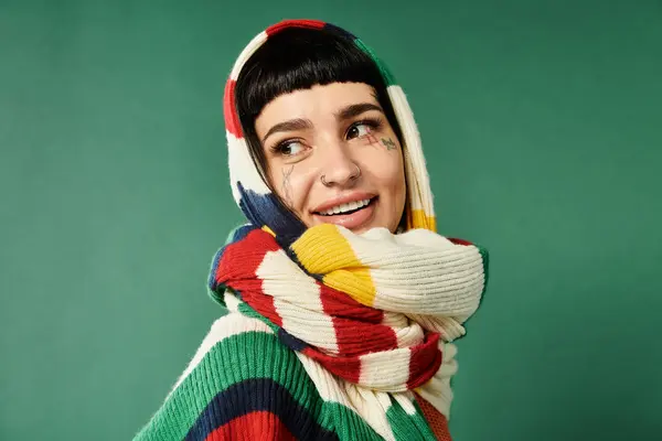 A young woman with short hair and tattoos smiles warmly while wearing a cozy sweater and scarf, set in a vibrant green studio. — Stock Photo