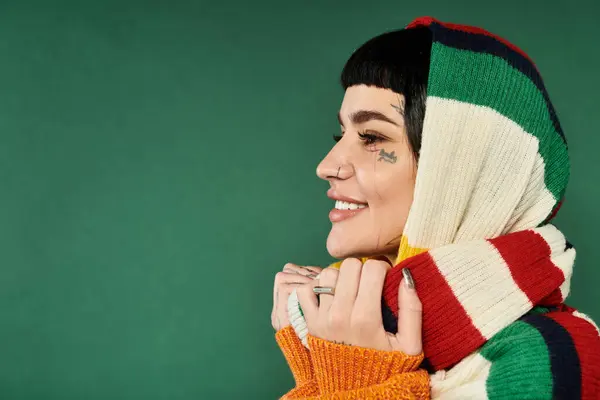 A young woman with short hair and tattoos smiles while wearing a vibrant, cozy sweater and scarf in a studio setting. — Stock Photo