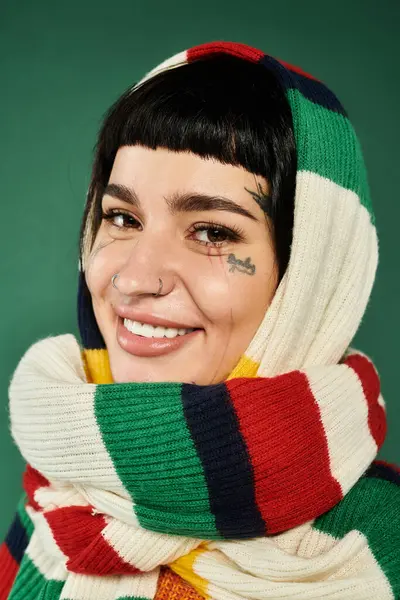 A young woman with tattoos and short hair poses cheerfully in a cozy sweater and scarf, surrounded by a green backdrop. — Stock Photo