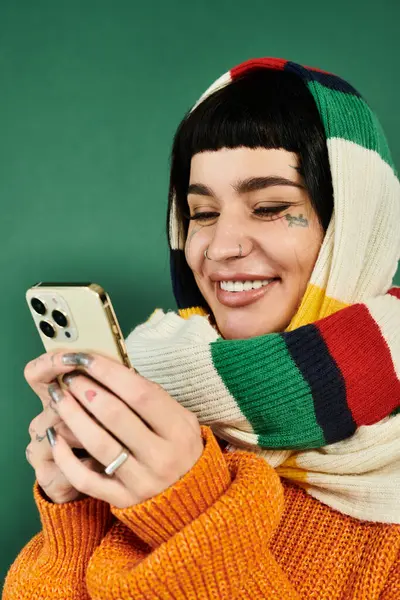 Молода жінка з коротким волоссям і татуюваннями посміхається, використовуючи свій телефон в барвистому светрі і шарфі на зеленому тлі. — стокове фото