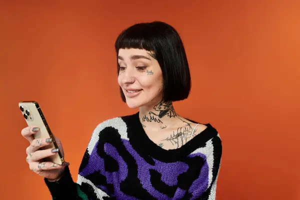A young woman with short hair and tattoos smiles while using her smartphone in a colorful setting. — Stock Photo