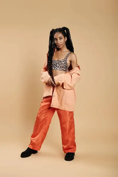 A confident young woman stands elegantly, flaunting her chic attire against a warm backdrop. — Stock Photo
