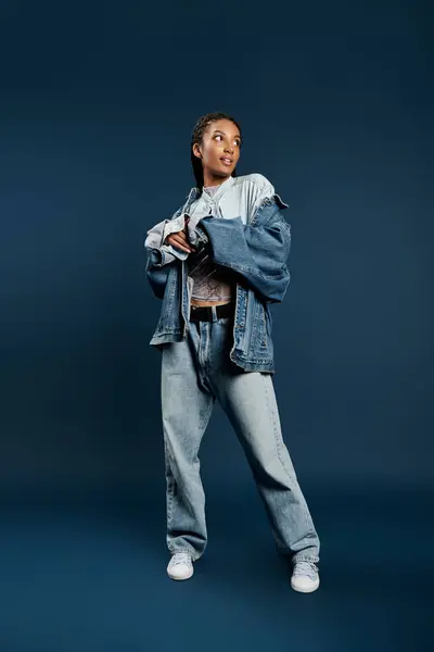 A beautiful young woman poses confidently in fashionable denim attire with a blue background. — Stock Photo