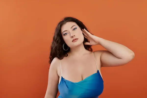 A young, beautiful plus size woman exudes confidence in her blue dress while posing on a bright orange background. — Stock Photo