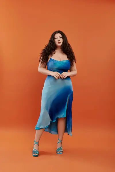 A beautiful young woman showcases her style in a blue dress, striking a pose on a bold orange background. — Stock Photo