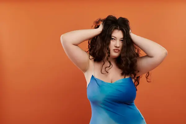 A vibrant young woman in a blue dress with makeup poses confidently on a bold orange background. — Stock Photo