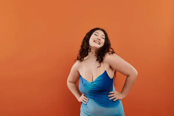 Una joven mujer irradia belleza y confianza en un vestido azul impresionante, situado sobre un fondo naranja cálido. - foto de stock