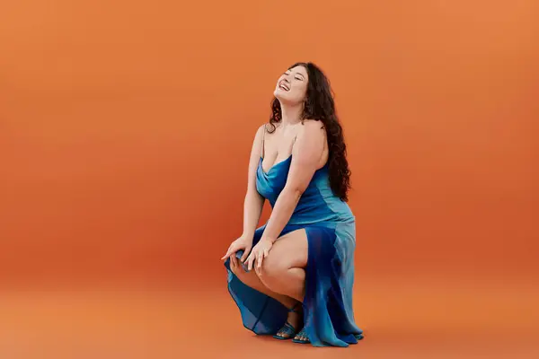 Une femme heureuse de taille plus dans une robe bleue s'agenouille et sourit vivement, mettant en valeur sa beauté sur orange vif. — Stock Photo