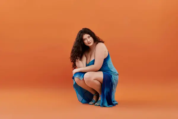 A confident plus size woman showcases her style in a stunning blue dress against a vibrant orange backdrop. — Stock Photo