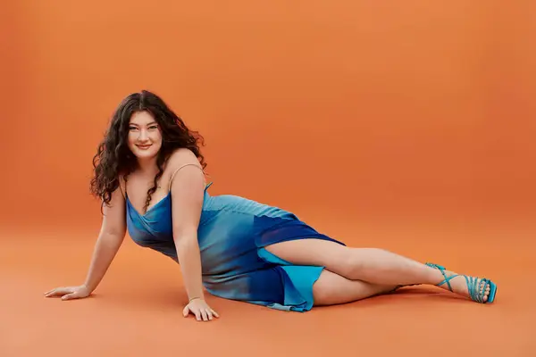Eine junge Frau stellt ihre Schönheit in einem blauen Kleid zur Schau und posiert selbstbewusst auf leuchtend orangefarbenem Hintergrund. — Stockfoto