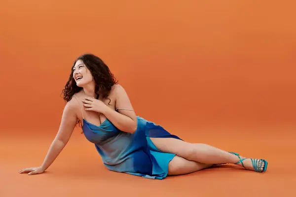 Eine selbstbewusste Plus-Size-Frau posiert glücklich in einem fließenden blauen Kleid vor leuchtend orangefarbenem Hintergrund. — Stockfoto