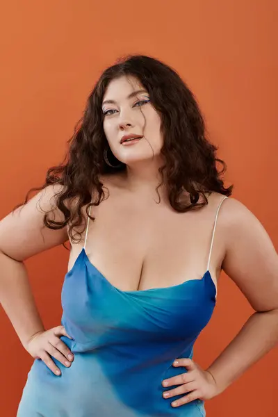 Яркая и уверенная в себе молодая женщина plus size демонстрирует свою красоту в потрясающем синем платье на оранжевом заднике. — стоковое фото