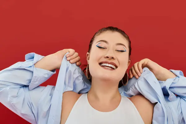 A joyful plus size woman showcases her vibrant makeup while playfully posing against a striking red backdrop. — Stock Photo