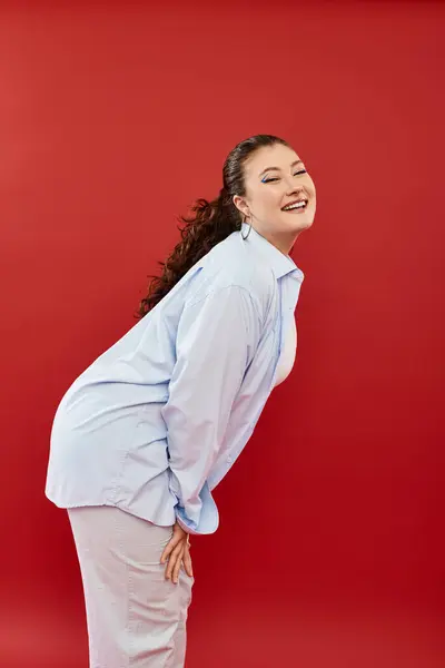 A joyful plus-size woman with striking makeup strikes a fun pose, radiating confidence on a bold red backdrop. — Stock Photo
