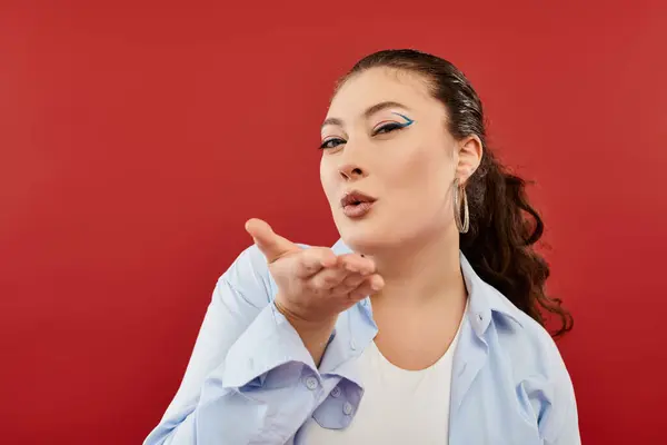 Una splendida donna plus size con trucco luminoso che soffia giocosamente un bacio in un vivace ambiente da studio. — Foto stock