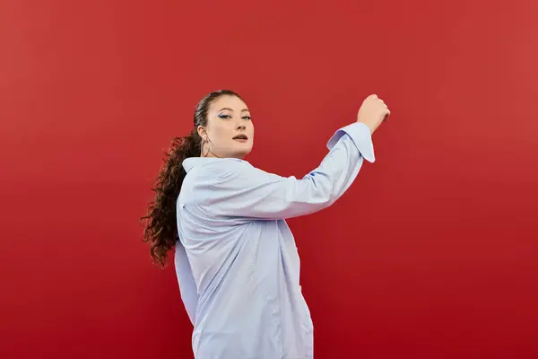 Una splendida donna plus size mette in mostra trucco luminoso mentre colpisce una posa di fronte a uno sfondo rosso vivido. — Foto stock