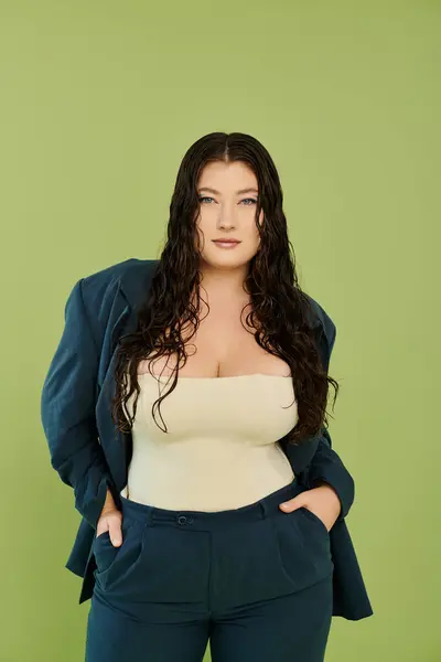 A young, beautiful plus size woman showcases her confidence in a chic suit while posing on a vibrant green background. — Stock Photo