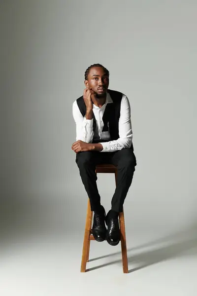 A young African American man sits thoughtfully on a stool, dressed in a sharp suit against a muted grey background. — Stock Photo