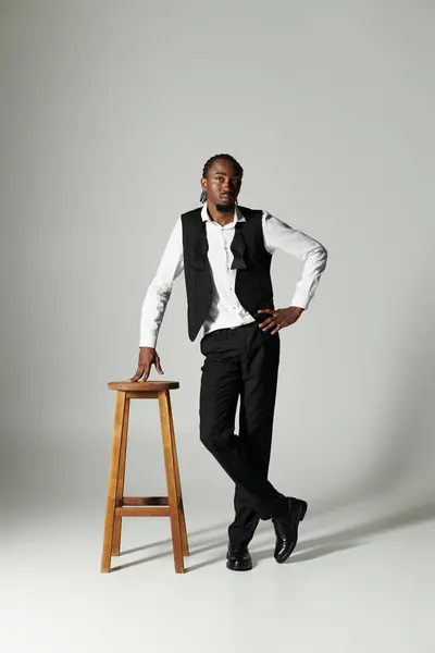 A young man in a suit stands confidently beside a wooden stool, striking a fashionable pose against grey — Stock Photo
