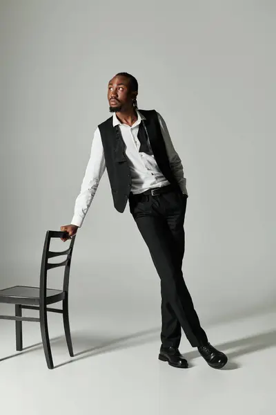 A suave young man stands elegantly with a chair, showcasing his chic suit and inner confidence in a grey setting. — Stock Photo