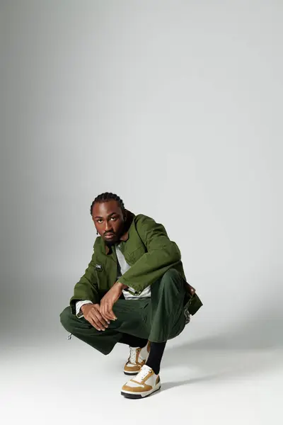 A fashionable young man in stylish green clothing crouches gracefully on a grey background, exuding confidence. — Stock Photo
