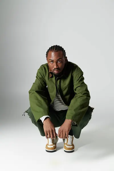 A young man wearing a fashionable green ensemble kneels and poses with style on a smooth gray background. — Stock Photo