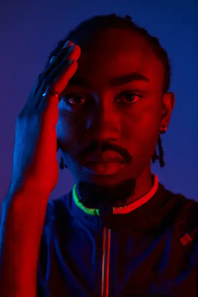A young African American man in trendy attire poses confidently with neon lights casting vibrant hues. — Stock Photo