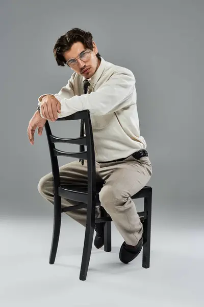 A young man dressed elegantly leans casually on a chair against a minimalist grey backdrop. — Stock Photo