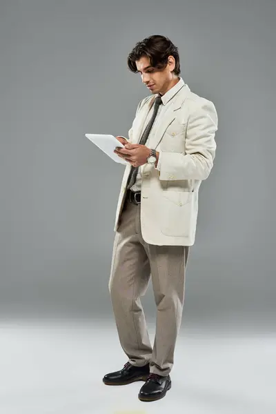 A young, handsome man in formal attire examines a tablet while posing against a sleek grey background. — Stock Photo