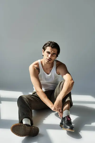 A young man sits casually on the floor, showcasing his tattoo while dressed in a tank top and stylish pants. — Stock Photo