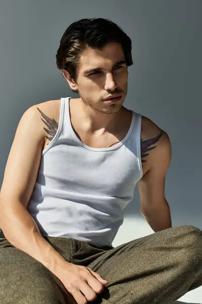 A young handsome man in a tank top showcases his tattoos while posing against a simple grey backdrop. — Stock Photo