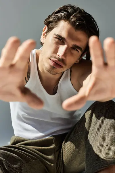 A handsome young man in a tank top playfully extends his arms forward, showcasing his tattoos on a grey background. — Stock Photo