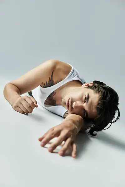 A young man in a tank top stretches out on a grey background, showcasing his tattoo, radiating relaxed charm. — Stock Photo