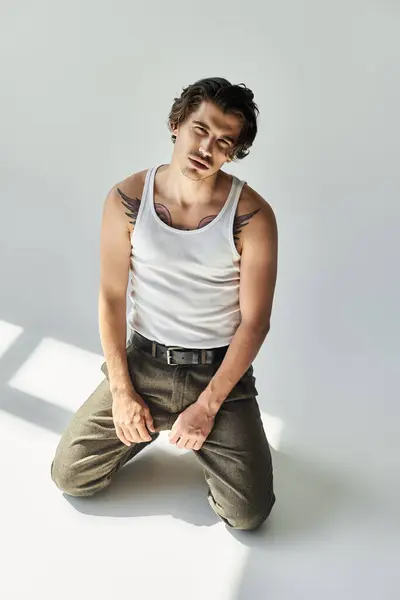 A young man with striking tattoos sits in a tank top, immersed in a thoughtful moment against a minimalist backdrop. — Stock Photo