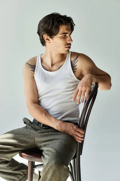 A young man in a white tank top sits elegantly on a chair, showcasing his tattoos against a plain grey background. — Stock Photo